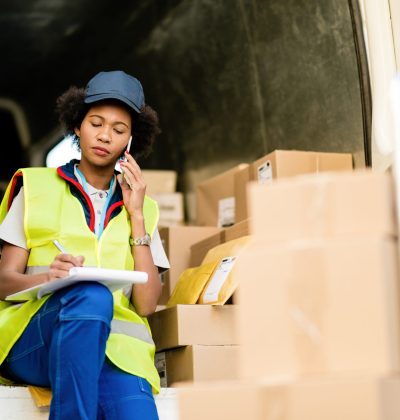 Female black courier sitting in delivery van and communicating on mobile phone while writing notes in clipboard.
