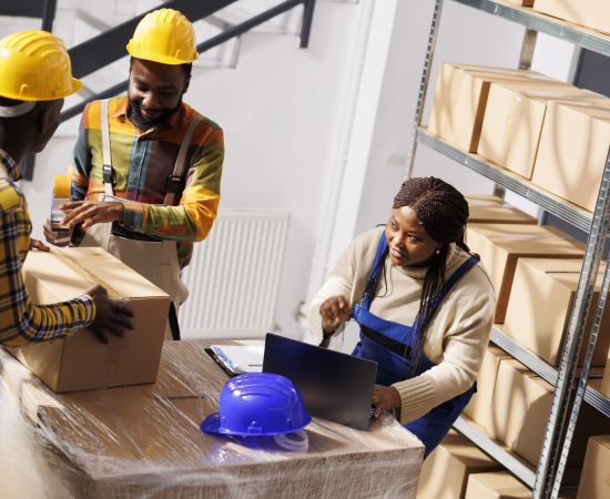 Warehouse manager checking orders list and coordinating parcels packing process. African american woman supervisor with laptop controlling freight cardboard boxes sealing and dispatching